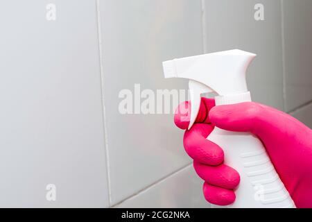 une main gantée vaporise un agent nettoyant d'une bouteille sur un mur en carreaux blancs Banque D'Images