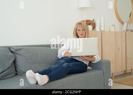 Femme d'âge mûr utilisant un ordinateur portable tout en étant assise sur un canapé confortable, à l'intérieur de la maison. Pleine longueur belle femelle shopping ou chat en ligne dans le réseau social, regarder le film, travail indépendant Banque D'Images
