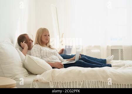 Portrait complet d'un couple d'âge mûr avec addiction numérique à la maison. Beau homme avec une tablette et belle femme d'âge moyen avec un ordinateur portable passer du temps ensemble tout en étant allongé dans le lit. Banque D'Images
