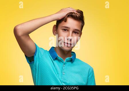 Le concept de l'adolescence et des émotions chez les adolescents. Un adolescent dans un t-shirt bleu, souriant sans aucun doute, égratignure sa tête. Arrière-plan jaune. C Banque D'Images