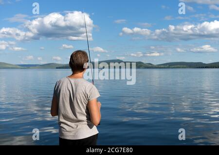 Une femme pêcheur se tient avec une canne à pêche et attrape des poissons contre un lac bleu et un ciel avec des nuages. Prise de vue lumineuse. Banque D'Images