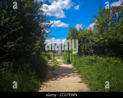 Ancienne clôture en métal fermée abandonnée avec porte tournante sur un sentier solitaire vide dans un parc sauvage vert avec des montagnes et un ciel bleu à l'arrière-plan. Banque D'Images