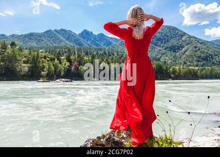 Vue arrière d'une femme blonde dans une longue robe rouge avec ses mains jetées derrière sa tête contre le fond de la rivière, les montagnes couvertes Banque D'Images