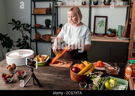 Contenu blonde femme mûre debout à la table de cuisine et de trancher la poire tout en enregistrant la vidéo de cuisine sur smartphone, vlog concept Banque D'Images