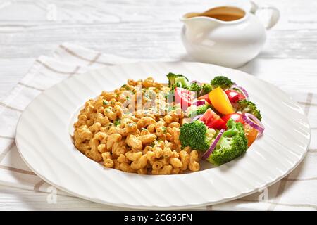 gros plan d'un mac vegan et du fromage avec une sauce à la levure et une salade de légumes sur une assiette blanche sur une table en bois blanche avec des couverts argentés, landsc Banque D'Images