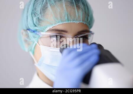 Femme médecin assistant de laboratoire dans les lunettes et le masque regarde à travers le microscope. Banque D'Images