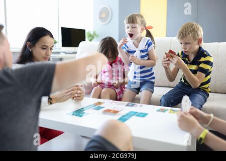 Les adultes et les enfants sont assis à la table et tiennent les cartes de jeu que la fille se leva et crie. Banque D'Images