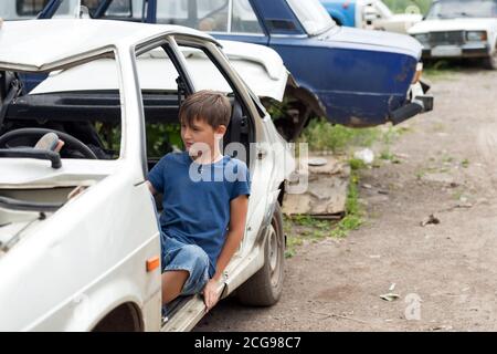 Un garçon de 11 ans est assis dans une voiture démontée dans un dépotoir de vieilles voitures abandonnées par une belle journée d'été. Banque D'Images