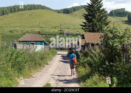 Un garçon de 12 ans, avec un sac à dos, marche le long d'une route de terre en passant devant une clôture en bois dans le village, par une journée ensoleillée d'été. Banque D'Images