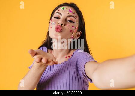 Image d'une femme heureuse avec des autocollants sur le visage soufflant de l'air baiser tout en prenant le selfie isolé sur le mur jaune Banque D'Images