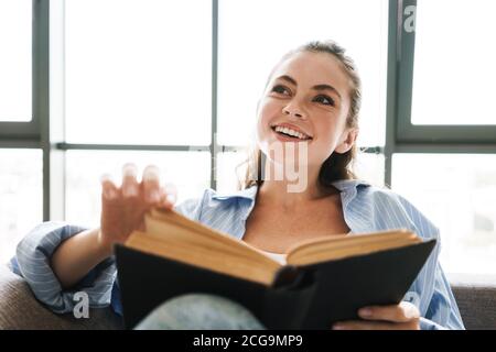 Photo d'une jeune fille heureuse rêvant à l'intérieur à la maison asseyez-vous sur le canapé pendant que vous lisez un livre Banque D'Images