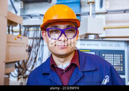 Jeune homme asiatique métallurgiste en casque orange et verres protecteurs bleus posé dans une usine de métallurgie du titane. Sur le fond du four sous vide. Banque D'Images