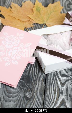 Guimauve dans une boîte cadeau. Attaché avec un ruban attaché à un noeud. À côté de la carte de vœux. Feuilles d'érable séchées tout autour. Sur les planches de pin peintes en noir an Banque D'Images