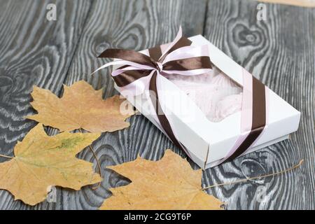 Zephyr dans une boîte cadeau. Attaché avec un ruban attaché à un noeud. Feuilles d'érable séchées tout autour. Sur les planches de pin peintes en noir et blanc. Banque D'Images