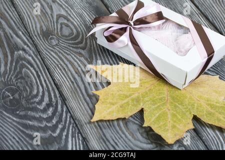 Zephyr dans une boîte cadeau. Attaché avec un ruban attaché à un noeud. Feuilles d'érable séchées tout autour. Sur les planches de pin peintes en noir et blanc. Banque D'Images