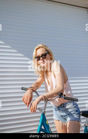 Fatigué après le vélo dans l'après-midi d'été chaud blonde meilleur femelle en short jeans et lunettes de vélo avec permanent ou d'un cycle qui pose sur bac blanc Banque D'Images