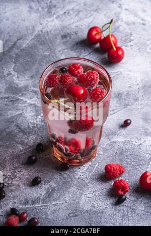 Boisson fraîche et froide à l'eau pétillante avec baies de cerisier, framboise et cassis en verre à facettes rouge sur fond de béton de pierre, boisson diététique d'été Banque D'Images