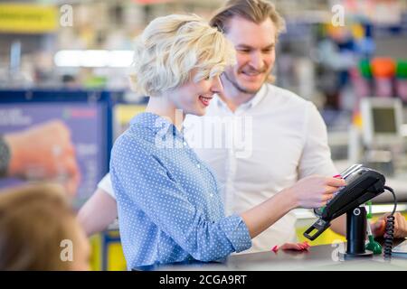 Une jeune femme blonde souriante se tenant dans un supermarché près du bureau de la caisse et payant avec une carte de crédit, tandis que son petit ami caucasien est à l'écoute du pur Banque D'Images