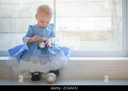 Une petite fille est assise sur le rebord de la fenêtre contre la fenêtre avec un décor hivernal. Enfant tient le jouet de l'arbre de Noël Banque D'Images