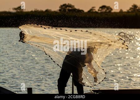 Un pêcheur portant une combinaison et des bottes ainsi qu'un chapeau de seau jette un filet moulé dans la mer depuis une jetée en bois au coucher du soleil. Il est rétroéclairé et voit Banque D'Images