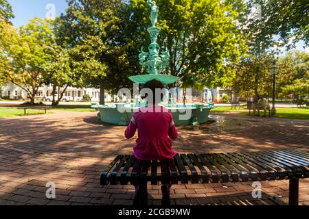 Une petite fille portant des vêtements décontractés roses est assise seule sur un banc de métal dans le parc Fountain de Chestertown, Maryland. En arrière-plan flou Banque D'Images