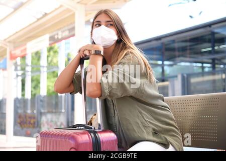 Jeune femme portant un masque, se reposant sur sa valise, regardant le train à une gare. Concept voyageur. Banque D'Images