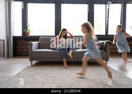 Jeune mère célibataire travaillant sur un ordinateur portable dans un loft assis sur un canapé pendant que ses filles courent autour d'elle et crient. Banque D'Images