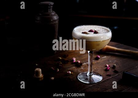 Sauer cocktail dans un grand verre de cocktail avec des stands de roses séchées sur le comptoir d'un bar sombre. Banque D'Images