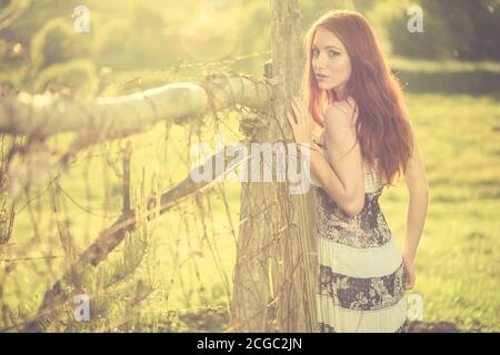 jeune femme à tête rouge portrait en robe vintage près de la vieille terre agricole clôture au coucher du soleil de printemps Banque D'Images