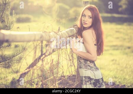 jeune femme à tête rouge portrait en robe vintage près de la vieille terre agricole clôture au coucher du soleil de printemps Banque D'Images