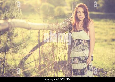 jeune femme à tête rouge portrait en robe vintage près de la vieille terre agricole clôture au coucher du soleil de printemps Banque D'Images