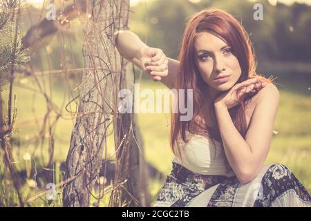 jeune femme à tête rouge portrait en robe vintage près de la vieille terre agricole clôture au coucher du soleil de printemps Banque D'Images