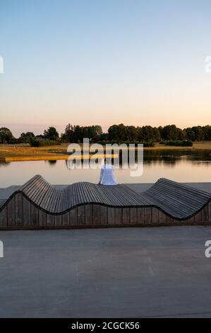 Une femme en chemise blanche assise sur un banc en bois vue sur un lac calme au coucher du soleil Banque D'Images