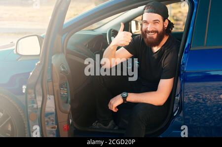 Photo d'un homme joyeux et barbu assis dans sa nouvelle voiture et montrant les pouces vers le haut. Banque D'Images