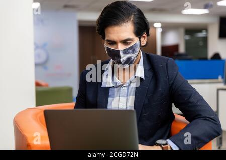 Portrait d'un jeune homme d'affaires indien réussi en costume sombre portant un masque de protection Covid et travaillant sur un ordinateur portable, environnement d'entreprise. Banque D'Images
