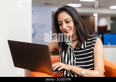 Portrait d'une femme d'affaires indienne prospère assise sur un canapé et travaillant sur un ordinateur portable, environnement d'entreprise. Banque D'Images
