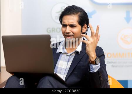 Portrait d'un jeune homme d'affaires indien en costume sombre, assis et travaillant sur un ordinateur portable, un environnement d'entreprise, un visage expressif. Banque D'Images