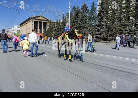 Un petit garçon se promette sur un cheval marron élégant conduit par un jeune homme le long de l'avenue de la ville de Krasnoyarsk pendant la célébration de la Seconde Guerre mondiale du jour de la victoire. Banque D'Images