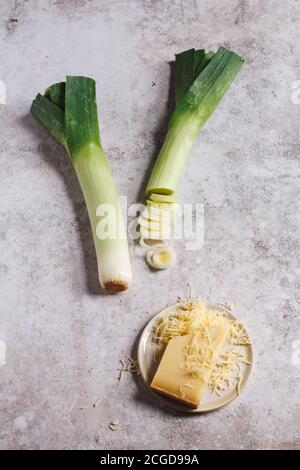 Poireaux frais et émincés avec une petite assiette de fromage français Comté râpé sur un plateau de table gris texturé. Banque D'Images