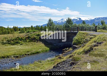 La rivière Merkjá passe sous un petit pont près du parking de Gluggafoss. Le glacier Eyjafjallajokull est visible dans les montagnes derrière les arbres. Banque D'Images