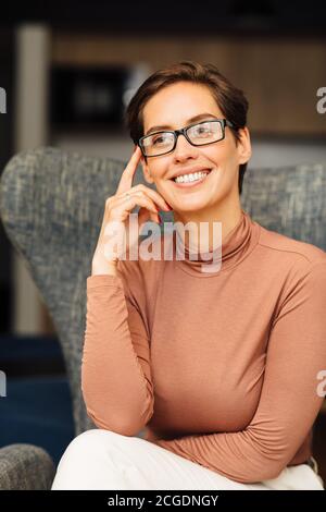 Belle femme souriante dans des lunettes de vue se détendre dans un fauteuil. Portrait d'une femme d'affaires vêtue de vêtements décontractés dans l'appartement. Banque D'Images