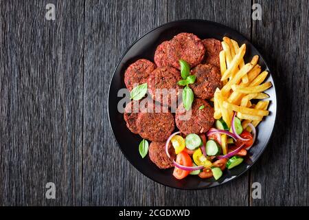 Patties de betteraves aux champignons et aux haricots noirs servis avec des frites et une salade de légumes de tomates, concombres, oignons rouges et basilic frais sur un p noir Banque D'Images