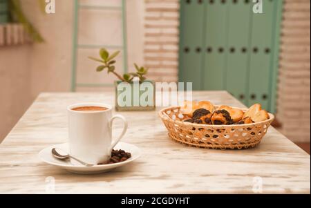 Une tasse de café sur une table en marbre, avec des pâtisseries dans un panier en osier et des plantes décoratives, et le fond de la maison méditerranéenne Banque D'Images