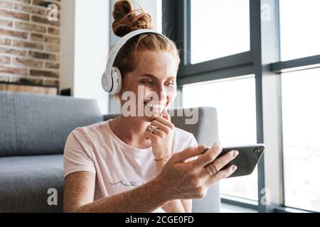 Image d'une femme souriante dans un casque et jouant à un jeu en ligne sur un téléphone portable, assis sur le sol à la maison Banque D'Images