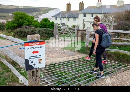 Les jeunes femmes marchent au-dessus de la grille de bétail à danger Cliff érosion panneau aux célèbres cottages de garde-côtes sur les falaises surplombant Cuckmere Haven Banque D'Images