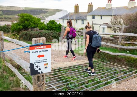 Les jeunes femmes marchent au-dessus de la grille de bétail à danger Cliff érosion panneau aux célèbres cottages de garde-côtes sur les falaises surplombant Cuckmere Haven Banque D'Images