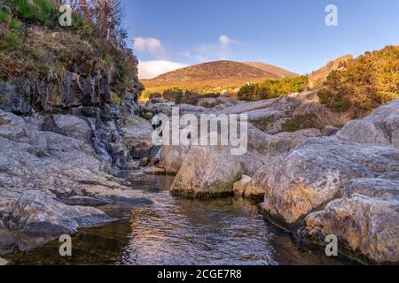 Ruisseau de montagne qui coule entre les rochers avec le sommet de montagne de Slieve Donard, mis en évidence par la lumière du soleil en arrière-plan, Mourne Mountains, Irlande du Nord Banque D'Images