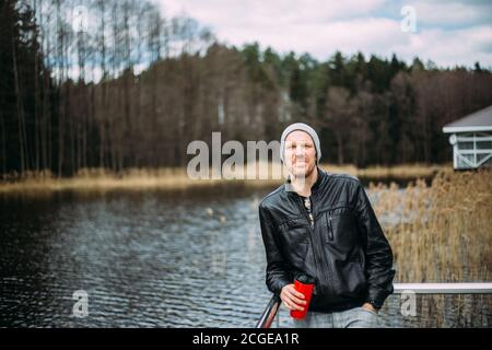 Homme pensif dans une veste en cuir et un chapeau se tient et pense à l'extérieur. Bon modèle mâle regardant pensive au lac. Banque D'Images
