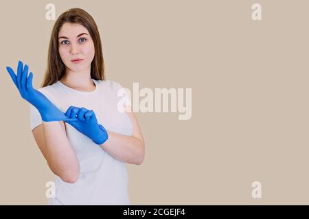 Une blonde aux cheveux longs posant, debout sur un fond clair avec un t-shirt blanc, mettant des gants médicaux blancs dans ses mains. Fournitures médicales. R Banque D'Images