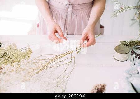 Mains d'une femme fleuriste au travail. Compositions sèches et bouquets de fleurs et de plantes pour l'intérieur. Fond rustique en bois blanc. Banque D'Images